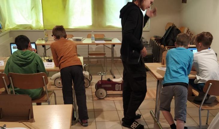 A coach works with a group of children at laptops to control model cars.